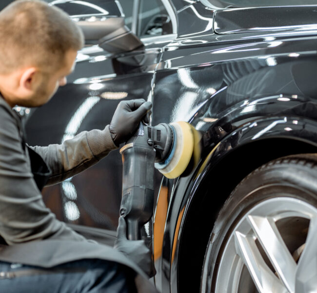 Worker polishing vehicle body with special grinder and wax from scratches at the car service station. Professional car detailing and maintenance concept